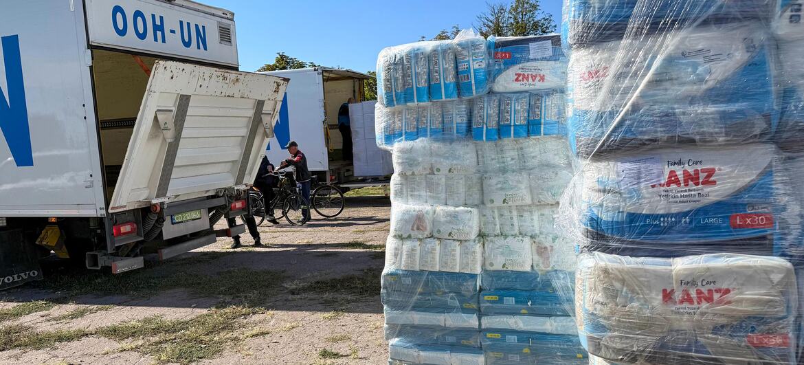 UN OHH-UN truck with open rear door unloads pallets of KANZ hygiene supplies and diapers at an outdoor distribution site in Kherson region; workers and bicycles stand nearby on a dirt ground under a clear blue sky.