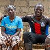 A Black African couple sits together on woven chairs in front of a mud-brick wall; the man has his arm around the woman and wears a red, white and blue sports shirt with rubber boots.