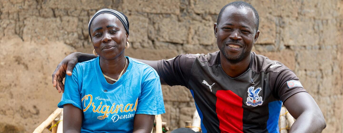 A Black African couple sits together on woven chairs in front of a mud-brick wall; the man has his arm around the woman and wears a red, white and blue sports shirt with rubber boots.