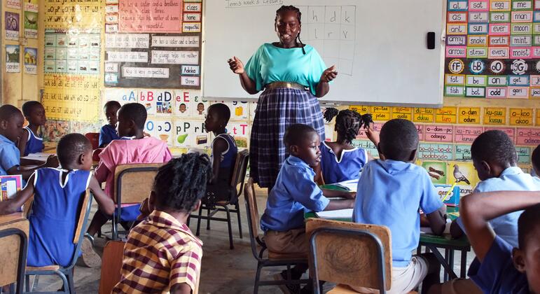 Dans une salle de classe d'une école primaire aux couleurs vives au Ghana, une enseignante se tient devant un tableau blanc et s'adresse à un groupe de jeunes élèves africains assis à leurs pupitres.