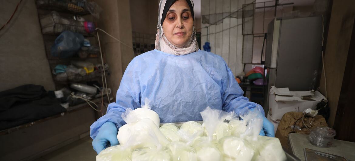 A woman wearing a headscarf and blue protective gown holds a metal tray of plastic-wrapped white food products in an indoor workshop.