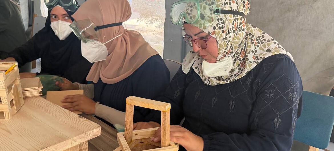 Three Arab women wearing hijabs, safety goggles and dust masks assemble a wooden stool frame at a workbench in a workshop.