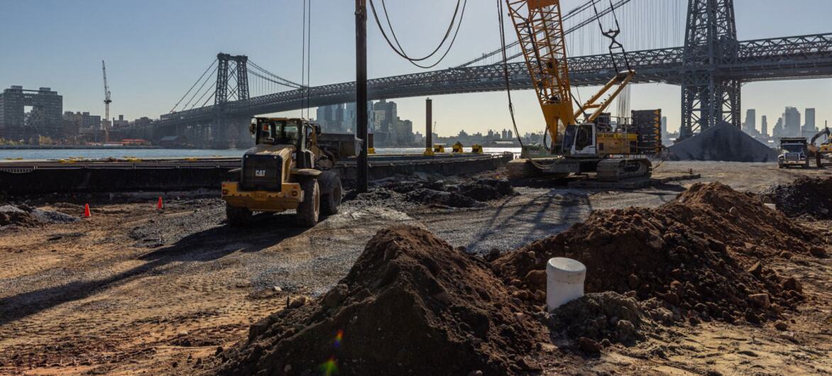 Heavy construction equipment including a yellow front loader and tower crane operates on a dirt site with piles of gravel in the foreground,