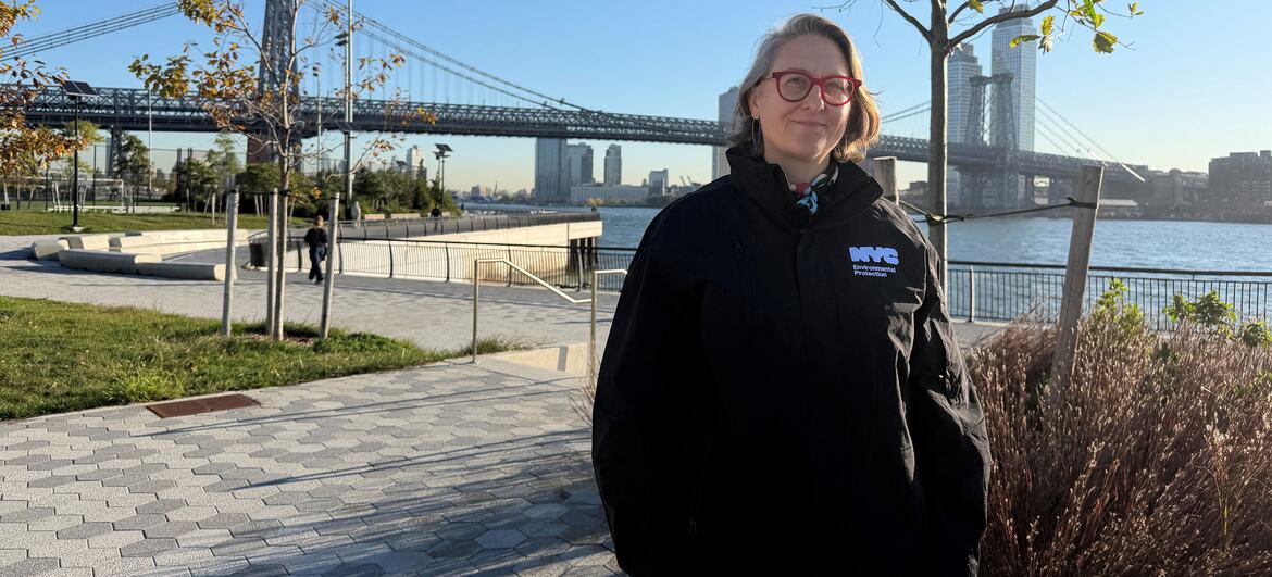 Kathryn Prybylski stands on a paved waterfront promenade at the East Side Coastal Resiliency project in Manhattan, with the Manhattan Bridge and East River behind her.