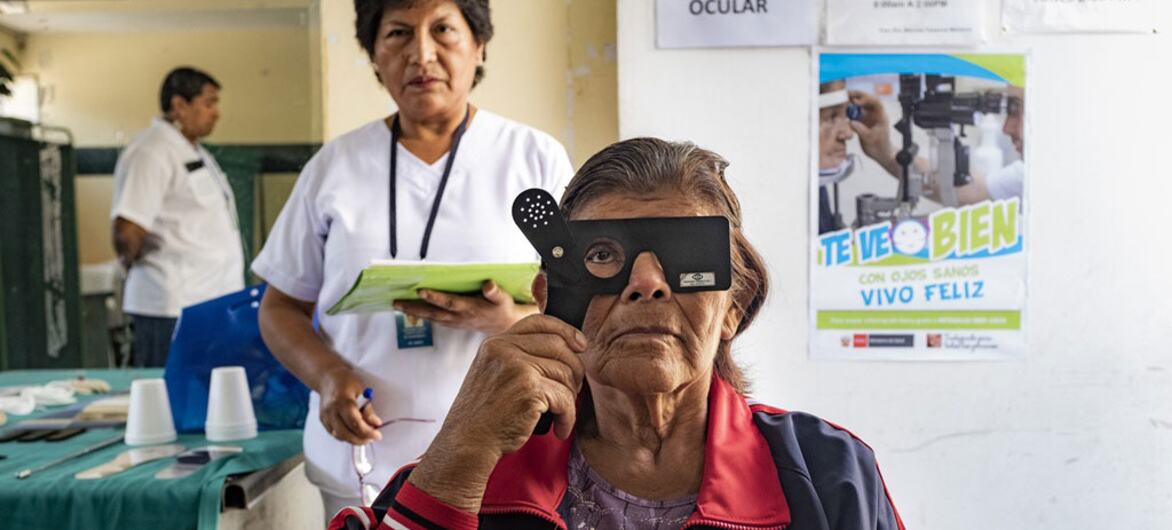 An elderly Indigenous woman in a Peruvian health clinic holds an examination instrument over one eye while a nurse stands behind her near wall signs for ophthalmic health.