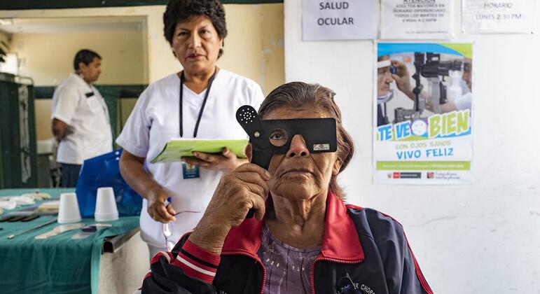 En una clínica de salud peruana, una anciana indígena sostiene un instrumento de exploración sobre un ojo mientras una enfermera se encuentra detrás de ella, cerca de los letreros de la pared que indican la ubicación de los dispositivos oftalmológicos.