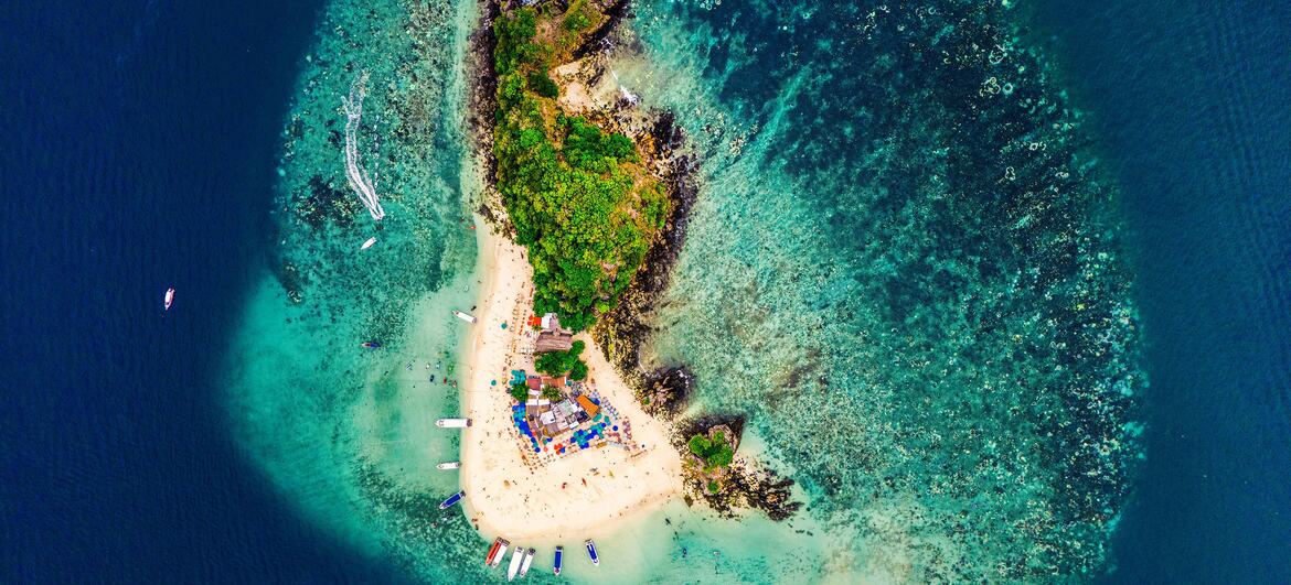 Vista aérea de uma pequena ilha tropical em Phuket, Tailândia, rodeada por águas rasas turquesa e recifes de coral, com barcos ancorados perto da praia de areia.