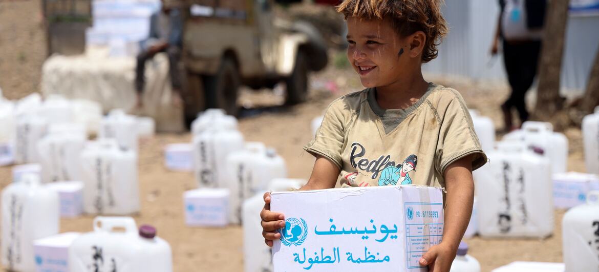 Hatem, de quatro anos, sorri enquanto segura uma caixa de kit de higiene da Unicef em um campo de deslocados internos em Jabal Habashi, Taiz, Iêmen, cercado por galões de água brancos.
