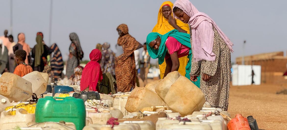 Black African women and children queue with jerrycans and collapsible water bladders for trucked chlorinated water at an outdoor distribution point in Al Obeid.