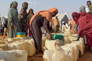 Black African women and children queue with jerrycans and collapsible water bladders for trucked chlorinated water at an outdoor distribution point in Al Obeid.