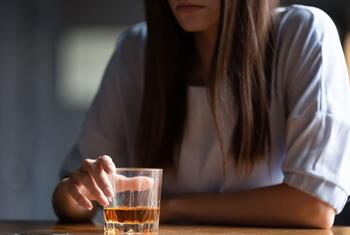 Gros plan sur une femme en chemisier blanc tendant la main vers un petit verre de liqueur ambrée posé sur un comptoir de bar en bois.
