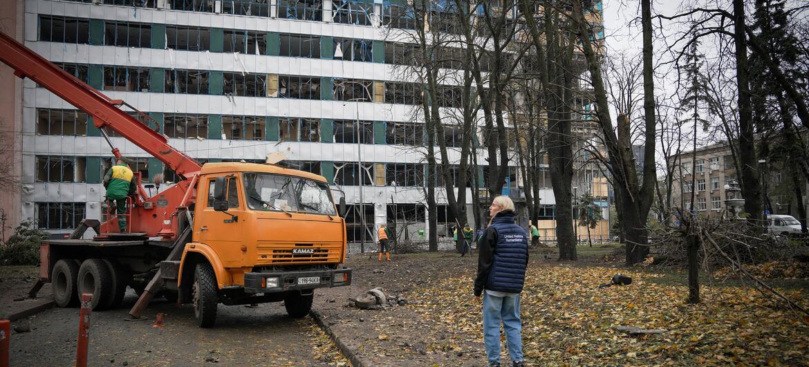 A UN OCHA staff member stands on a leaf-covered path beside an orange crane truck as workers work near a multi-storey building with shattered windows after a missile strike in Kyiv.