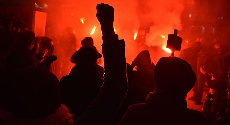 Foule en silhouette, poing levé, devant de la fumée rouge et des flammes lors d'un rassemblement nocturne dans la rue.