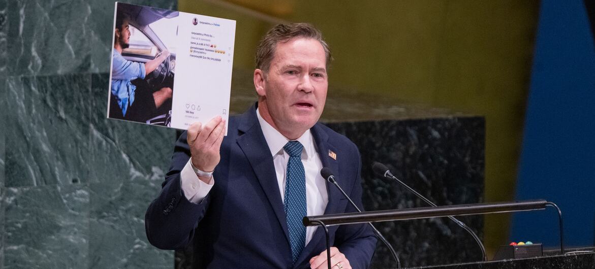A male white diplomat in a dark suit speaks at a United Nations General Assembly podium while holding up a printed social media post.
