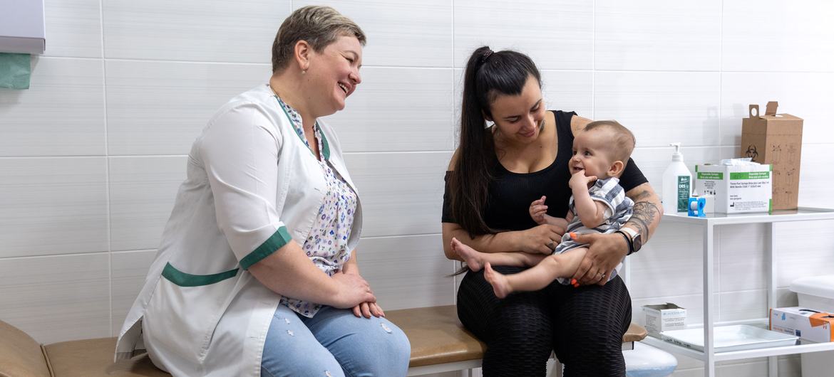 A smiling nurse and a mother holding an eight-month-old baby at a health center in Kyiv, Ukraine, preparing for vaccination.