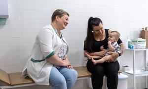 A smiling nurse and a mother holding an eight-month-old baby at a health center in Kyiv, Ukraine, preparing for vaccination.