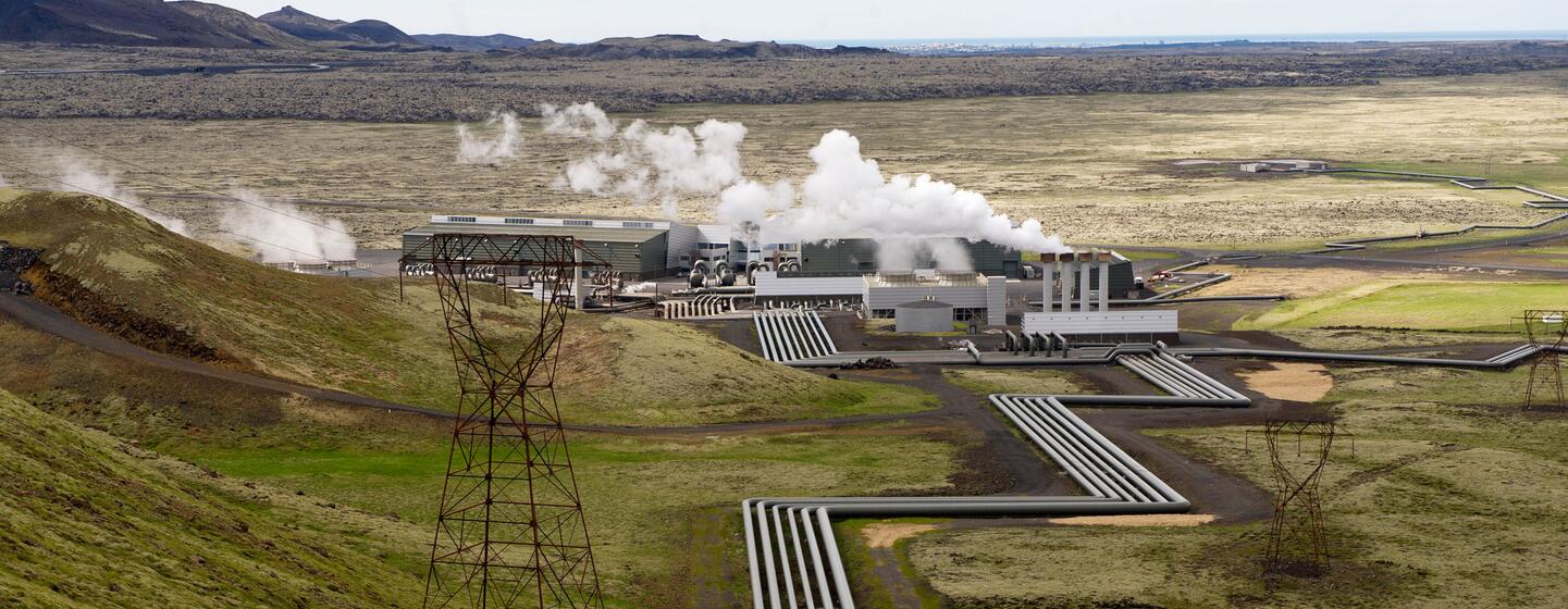 Una toma amplia de la central geotérmica de Hellisheiði en Islandia, que muestra el vapor que se eleva desde la instalación y grandes tuberías que serpentean por el paisaje verde y montañoso.