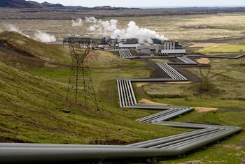 Una toma amplia de la central geotérmica de Hellisheiði en Islandia, que muestra el vapor que se eleva desde la instalación y grandes tuberías que serpentean por el paisaje verde y montañoso.