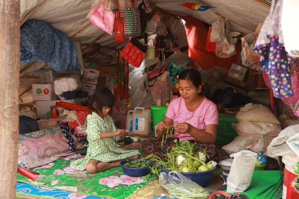 Familia moja ya wahamiaji kutoka Myanmar wanakimbilia ndani ya hema la muda huko Sagaing kufuatia tetemeko la ardhi la Machi 2025. Mwanamke wa Asia na msichana mdogo wanaonyeshwa wakiandaa mboga kwenye godoro ndani ya makao yaliyojaa fujo.