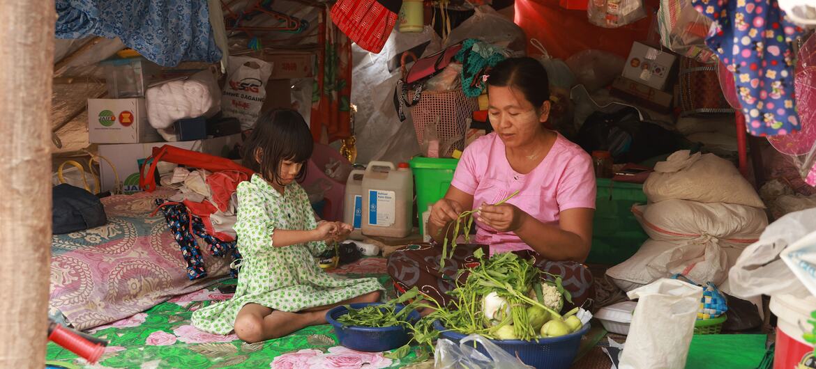 Uma família deslocada de Mianmar abriga-se dentro de uma tenda improvisada em Sagaing após o terremoto de março de 2025. Uma mulher asiática e uma menina são mostradas preparando legumes em um tapete dentro do abrigo desorganizado.