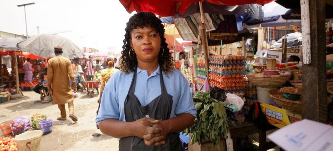 Uma vendedora de mercado está em sua barraca na Nigéria, em meio a um cenário de atividade comercial, ilustrando o impacto do aumento dos preços dos alimentos e da fome.