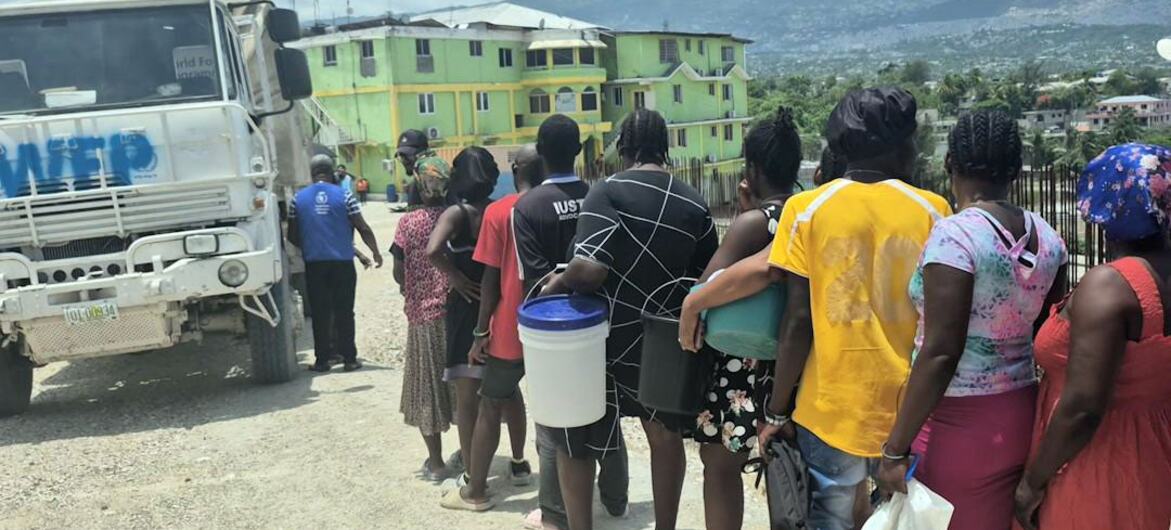 A line of people waiting for aid in front of a truck marked with 'WFP'. The individuals are standing on a gravel surface, with residential buildings in the background and cloudy skies above.