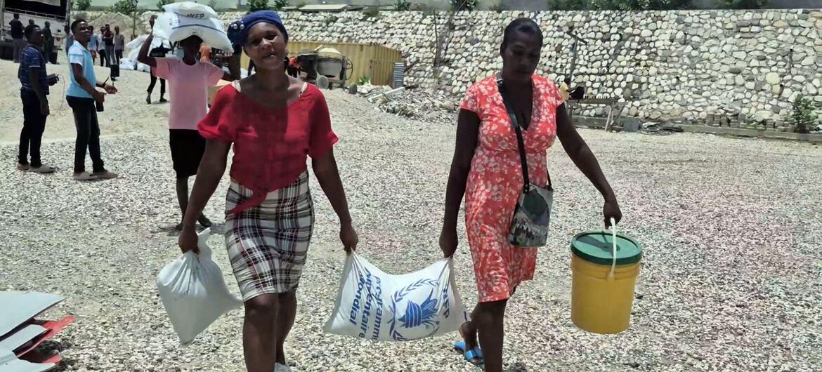 Displaced people in Haiti walk on a gravel road carrying food aid bags and a bucket.