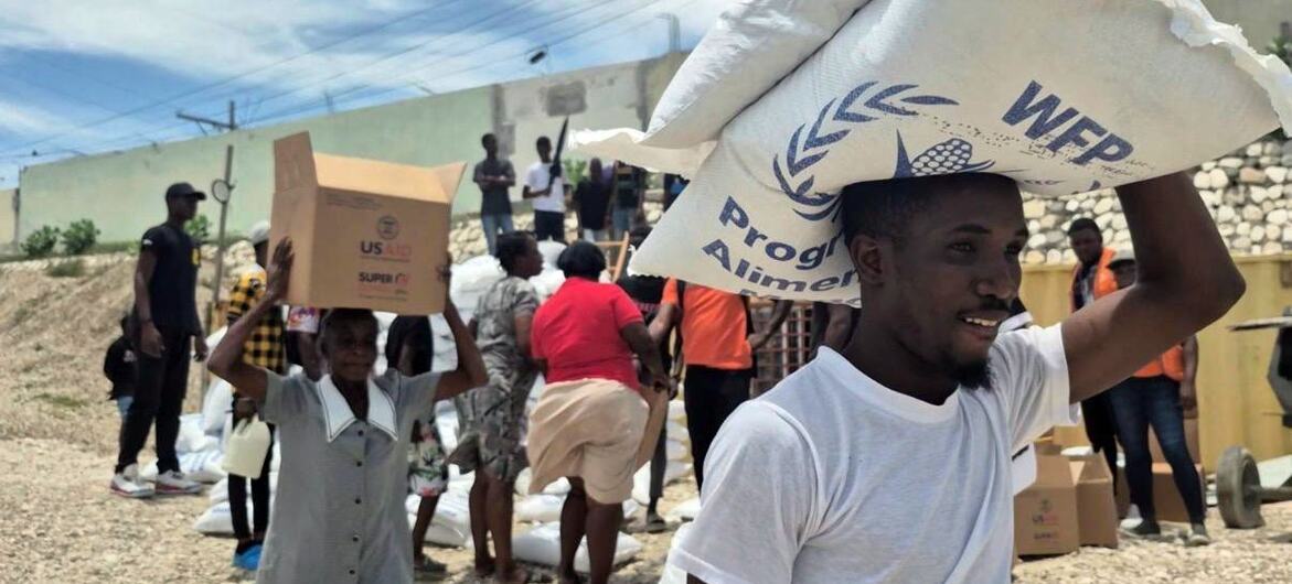 Residents of Cité Soleil carry WFP food aid parcels through Drouillard in Port-au-Prince, Haiti. A man in the foreground balances a large sack of rice on his head, while a woman carries a box of food assistance.