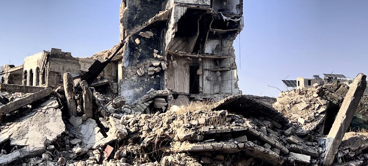 A heavily damaged multi-story building stands amidst a large pile of rubble and concrete debris under a clear sky.