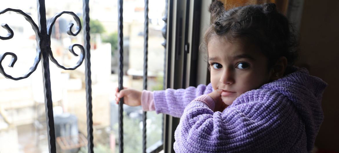 A young Palestinian girl in a purple sweater looks out a barred window from inside her home.