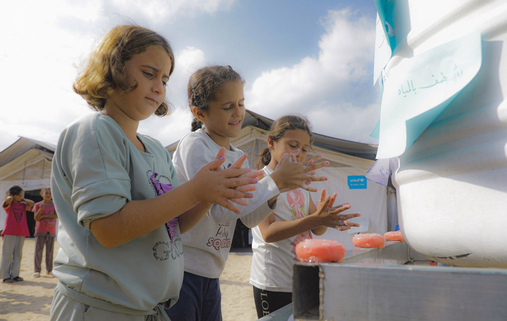 Três meninas lavam as mãos em um dispensador de sabão em um centro de ajuda humanitária em Gaza.