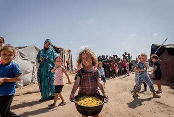 A smiling young girl, Shereen, carries a large pot of rice and chickpeas in Al-Mawasi, Khan Younis, while other children and adults stand near tents in the background.