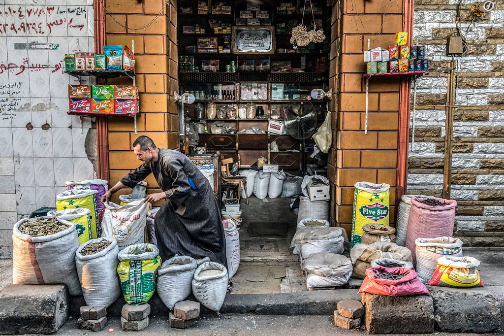 An Egyptian man in traditional clothing sorts through large sacks of grain and produce in the doorway of a small shop in Cairo.