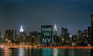 The United Nations Secretariat building illuminated with 'THANK YOU NY' against the New York City skyline at night, viewed from the East River.