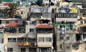 سلوان، مغربی کنارے میں کثرت سے بھری ہوئی عمارتوں کا فضائی نظارہ، جس میں بڑی آنکھوں کی دیواریں اور اسرائیلی جھنڈے نظر آتے ہیں، جس سے پڑوس کے پیچیدہ سیاسی اور سماجی مناظر پر روشنی ڈالی جاتی ہے۔