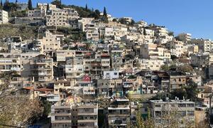 Una vista panorámica del barrio de Silwan en Jerusalén Oriental, que muestra edificios densamente llenos en una ladera de colina bajo un cielo azul claro. Algunas estructuras parecen dañadas o en construcción, lo que refleja el desplazamiento continuo y la expansión de los asentamientos.