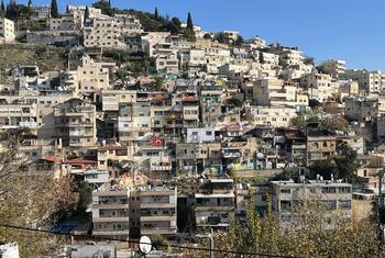 A panoramic view of the Silwan neighborhood in East Jerusalem, showing densely packed buildings on a hillside under a clear blue sky. Some structures appear damaged or under construction, reflecting ongoing displacement and settlement expansion.