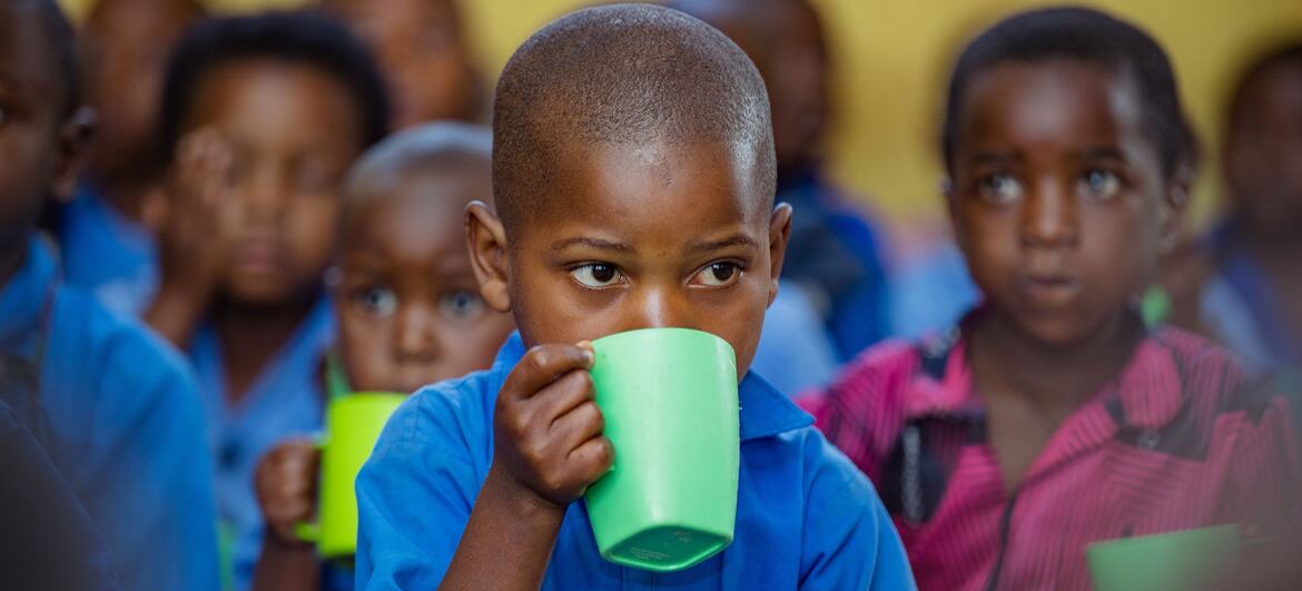A young child in a blue shirt drinks from a green cup in a classroom setting, surrounded by other children. This image captures a moment from a UNICEF-supported early childhood development initiative in Burera District, Rwanda.