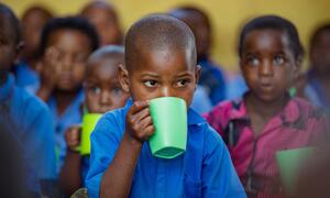 A young child in a blue shirt drinks from a green cup in a classroom setting, surrounded by other children. This image captures a moment from a UNICEF-supported early childhood development initiative in Burera District, Rwanda.