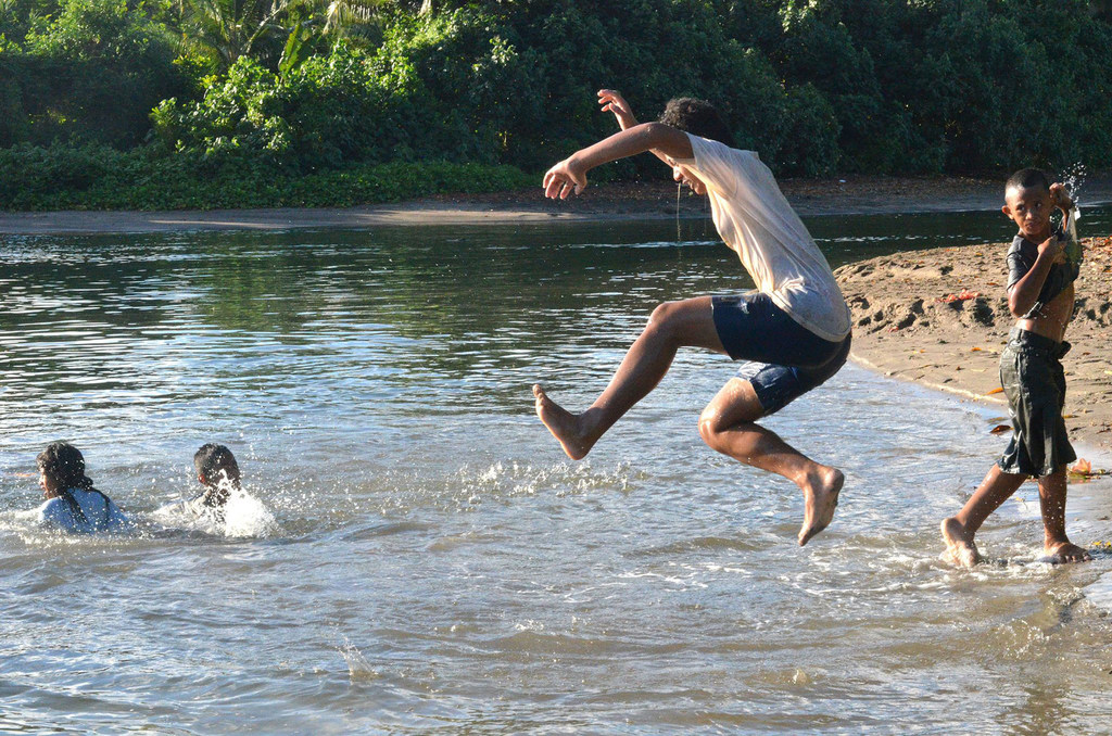 Niños jugando en un río en Samoa, con uno saltando al agua y otros nadando cerca.