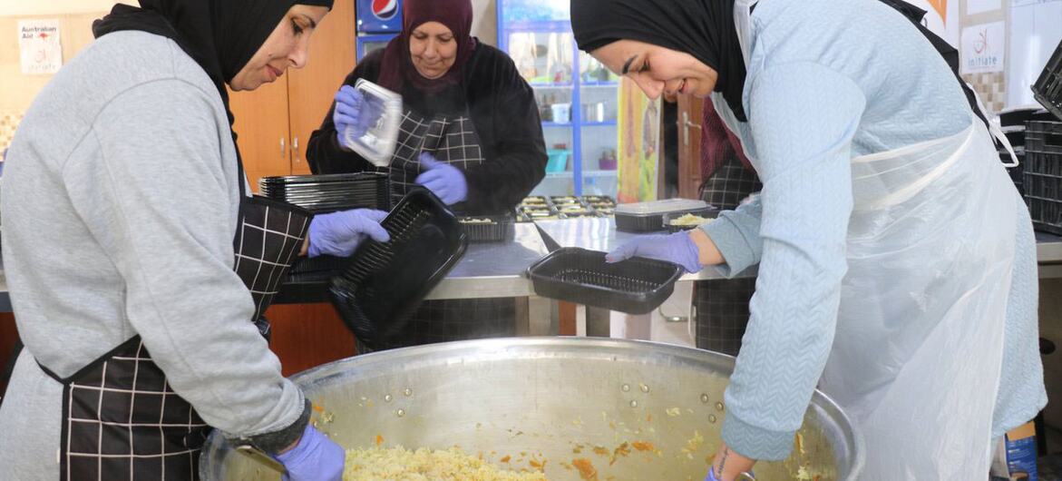 Tres mujeres en una cocina sirven arroz de una olla grande en contenedores. Llevan hiyab, delantal y guantes como parte de la iniciativa Semillas de Esperanza de ONU Mujeres en Tiro, Líbano.