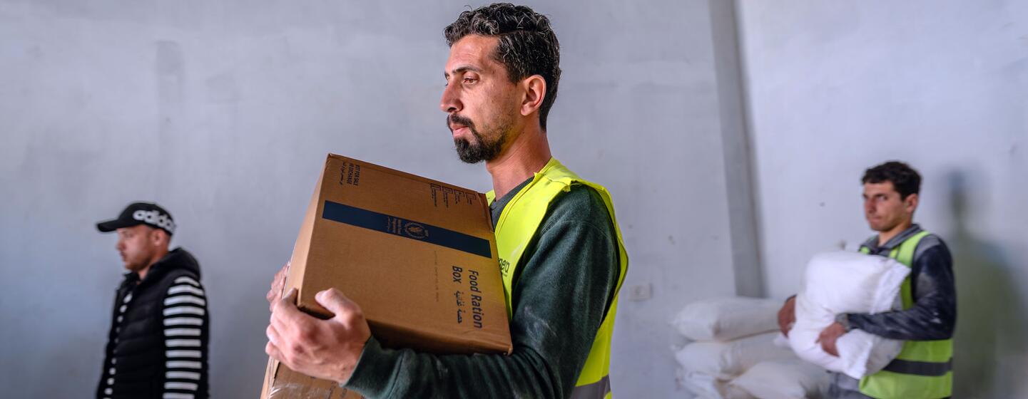 Trois hommes dans un entrepôt distribuent de l'aide alimentaire à Deir al-Balah, à Gaza. Un homme vêtu d'un gilet jaune porte une boîte en carton portant l'étiquette « Food Ration Box », tandis qu'un autre porte un grand sac blanc. L'aide, composée de biscuits à haute énergie du PAM, a été donnée par le gouvernement indonésien.