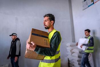 Three men in a warehouse distribute food aid in Deir al-Balah, Gaza. One man in a yellow vest carries a cardboard box labeled 'Food Ration Box', while another carries a large white sack. The aid, consisting of WFP high-energy biscuits, was donated by the Government of Indonesia.