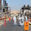 Workers in white hazmat suits walk through a designated warm zone at an industrial facility, with a security guard nearby.