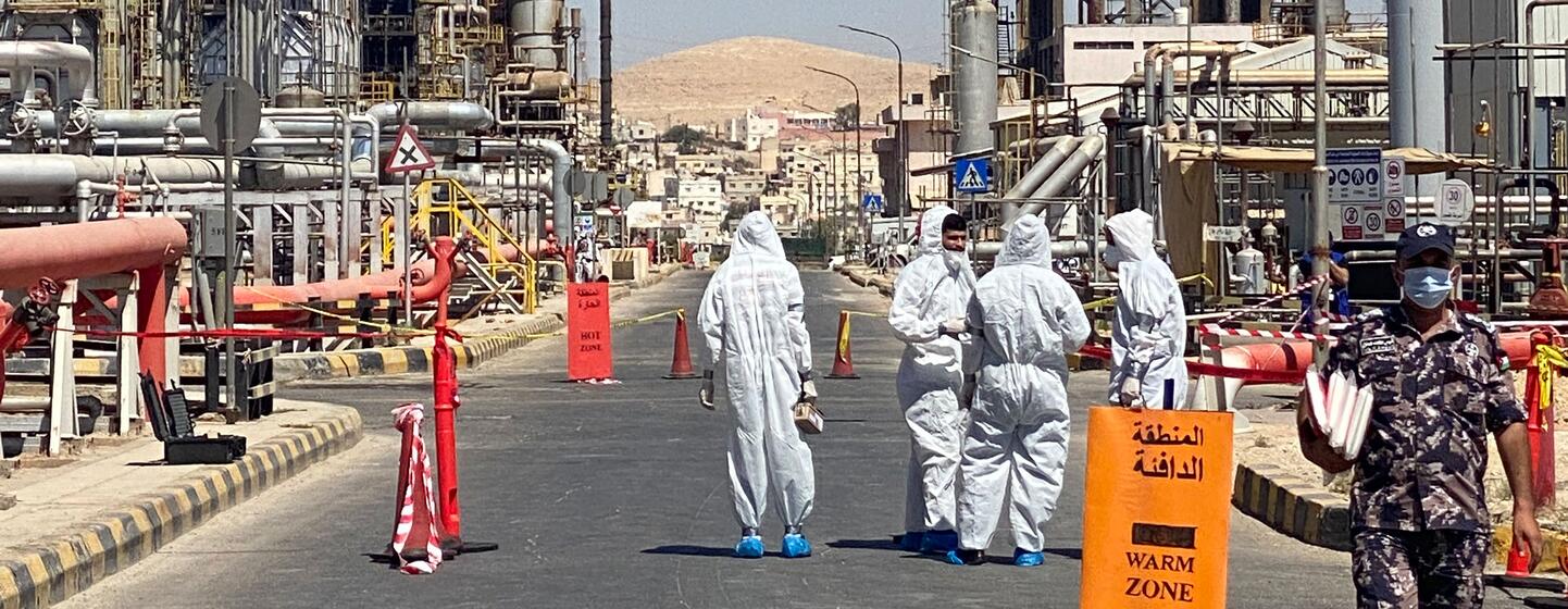 Workers in white hazmat suits walk through a designated warm zone at an industrial facility, with a security guard nearby.