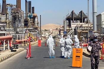 Workers in white hazmat suits walk through a designated warm zone at an industrial facility, with a security guard nearby.