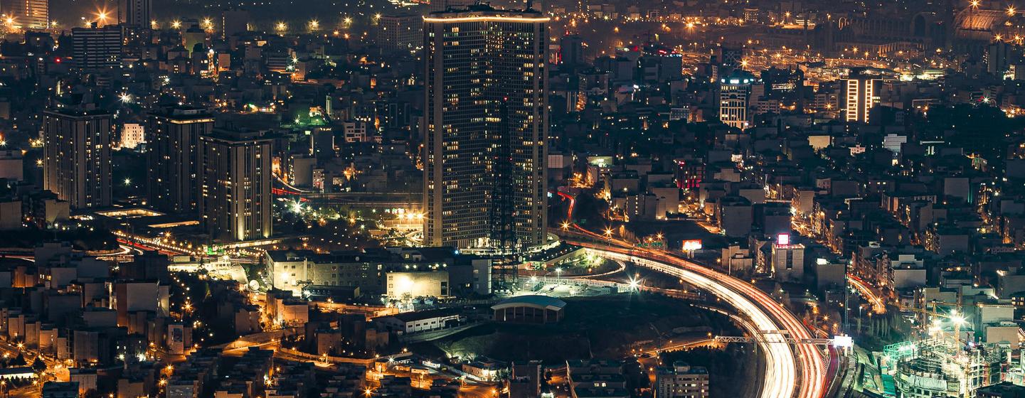 Une vue nocturne aérienne de Téhéran, en Iran, montrant un paysage urbain dynamique avec des immeubles de grande hauteur éclairés, des quartiers urbains denses et des rayures de lumière provenant de véhicules en mouvement sur les routes principales. L'image capture l'énergie dynamique et l'architecture moderne de la capitale sous le ciel nocturne.