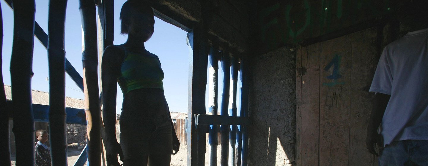 Uma jovem está parada na entrada de uma construção simples no Haiti, com um homem parcialmente visível ao lado.