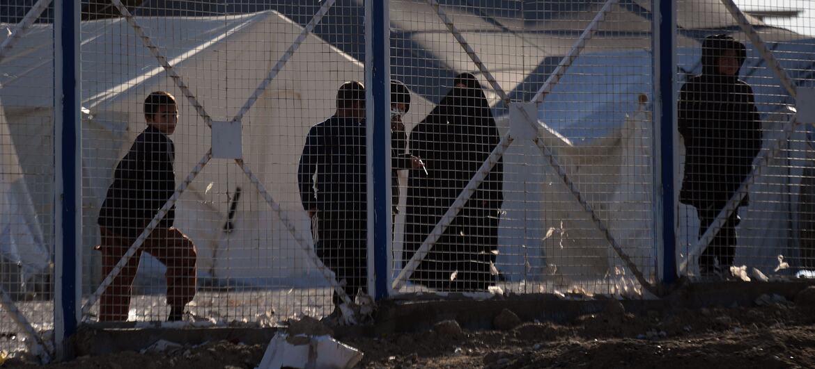 Silhouettes of people, including women and children, standing behind a metal fence at the Al-Hol Camp in Syria, with white tents visible in the background.