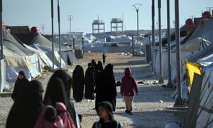 Women and children walk through the Al-Hol Camp in Syria, a displacement site housing over 30,000 people, with tents and infrastructure visible under a clear sky.